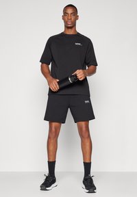 Férfi áll, fekete sportöltözetben, kezében fekete vízflakont tart, egyszínű világos háttér előtt.