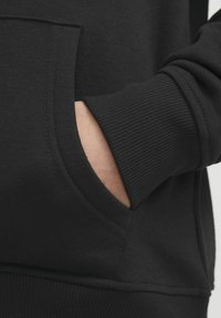 Sudadera negra con un bolsillo frontal, puños acanalados y una textura de tela suave, que destaca por sus líneas limpias y diseño minimalista.