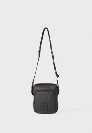 Sac bandoulière noir en matériau lisse, doté d'une poche avant, d'une fermeture éclair, d'une sangle réglable et d'un logo de marque en relief.