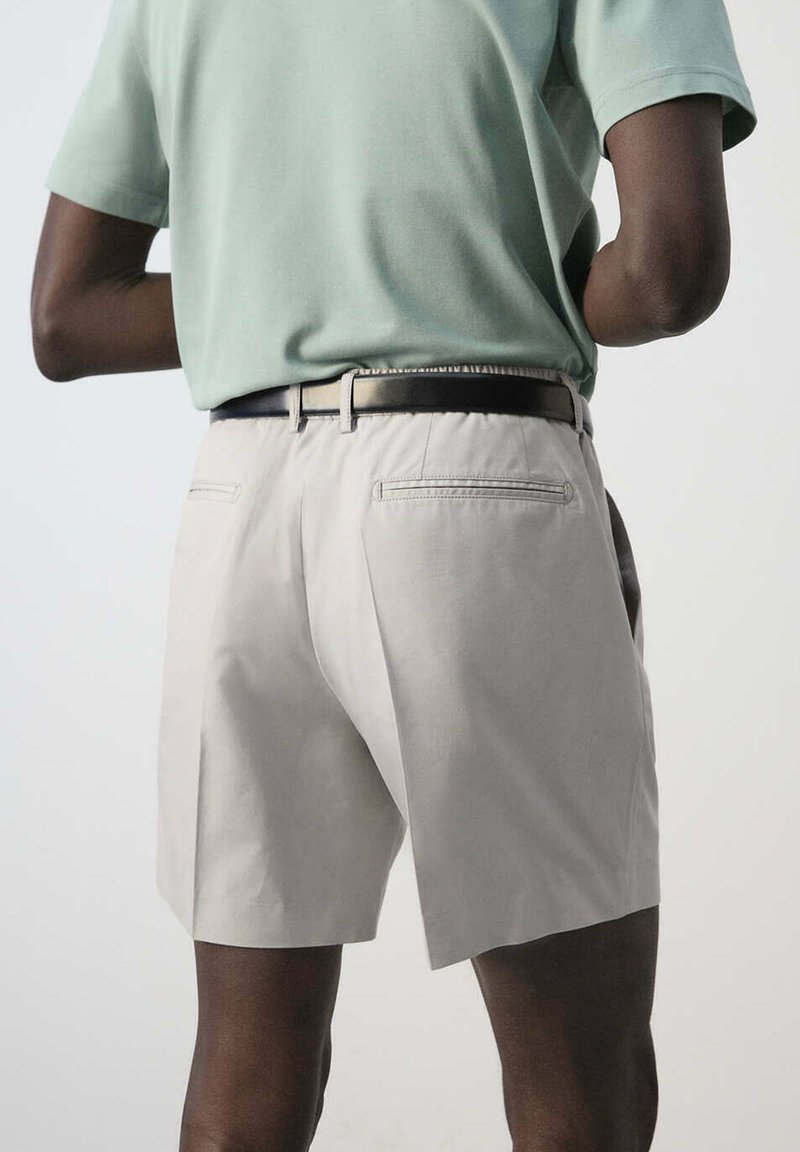 Man wearing light green short-sleeve shirt tucked into beige shorts with a black belt, shown from the back against a plain background.