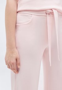 Κοντινό πλάνο ενός ατόμου που φοράει ανοιχτό ροζ παντελόνι με κορδόνι στη μέση και μπροστινή τσέπη, με το αριστερό του χέρι χαλαρά στο πλάι.