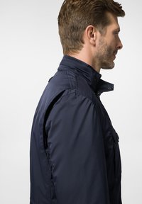 Granatowa kurtka z wysokim kołnierzem, zapinana na klapę i posiadająca boczne kieszenie. Wykonana z gładkiego, wodoodpornego materiału i o dopasowanym kroju.