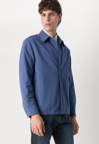 Blå jakke lavet af glat stof, med spids krave, knaplukning og sidelommer. Båret over en stribet skjorte og denim.
