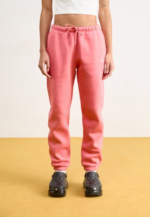 Pink sweatpants with a drawstring waist, featuring a small white logo. Paired with black clogs, set against a yellow floor.