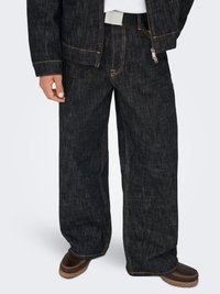 Zwarte spijkerbroek met een losse pasvorm, zichtbare stiksel details en een ritssluiting aan de zijkant. Gecombineerd met bruine schoenen.