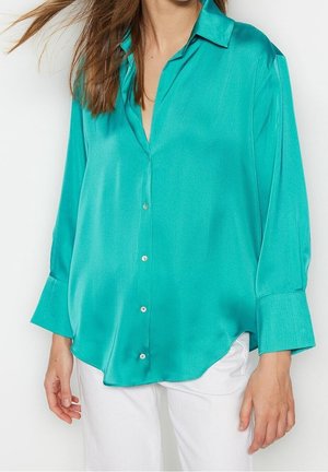 Kvinde iført en løs teal satin skjorte med knapper og brede manchetter samt hvide bukser, foran en ensfarvet lys baggrund.
