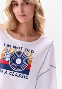 T-shirt graphique blanc avec une illustration de disque vinyle rétro et le texte "JE NE SUIS PAS VIEUX, JE SUIS UN CLASSIQUE." Détail de couture bleue contrastée.