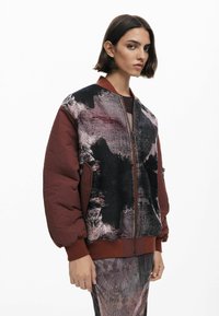 Quiltad bomberjacka i burgundy med ett texturerat svart och grått abstrakt mönster. Har ribbade ärmslut och en framficka med blixtlås.
