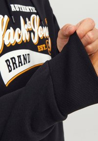 Schwarzer Sweatshirt mit einem grafischen Design in Weiß und Orange. Der Bund hat eine gerippte Textur, die eine Nahaufnahme der Details des Stoffs zeigt.