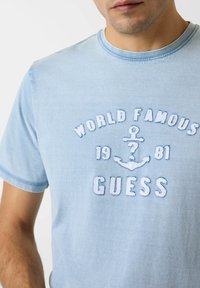 T-shirt en coton bleu clair avec texte brodé "WORLD FAMOUS 1981 GUESS" et un motif d'ancre, col rond et manches courtes.