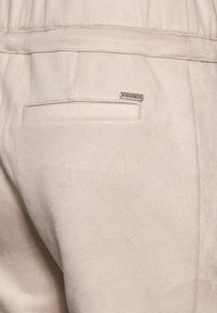 Beige broek met een gladde textuur, voorzien van een achterzak en een klein metalen label met de merknaam dat in de stof is genaaid.
