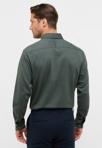 Camicia a maniche lunghe di un verde scuro con una texture liscia, polsini con bottoni e colletto classico; abbinata a pantaloni blu navy.