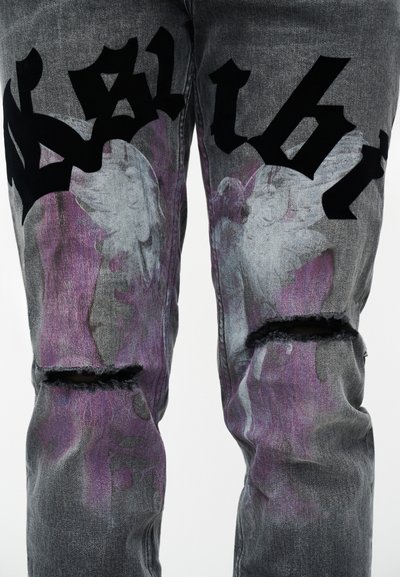 Grijze versleten spijkerbroek met gescheurde knieën, paars-witte verfachtige patronen en gedurfde zwarte gotische letters over de dijen.