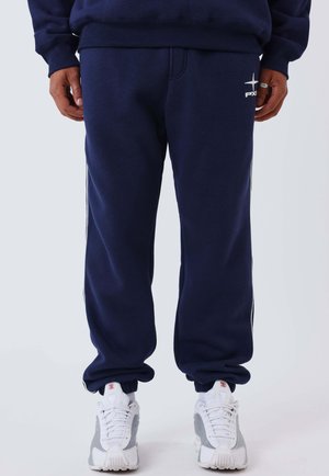 Uomo che indossa pantaloni della tuta blu navy con logo bianco sulla coscia sinistra, abbinati a scarpe da ginnastica bianche e grigie, in piedi contro uno sfondo semplice.