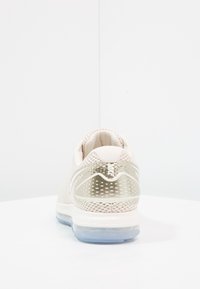 Baskets Nike beiges vues de l'arrière, avec un accent de talon métallique et une semelle bleue transparente sur une surface blanche.