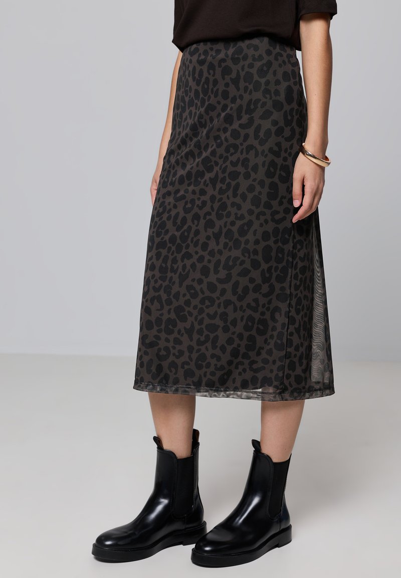 Jupe midi en imprimé léopard noir en tissu transparent, avec une texture lisse. Associée à des bottines noires brillantes et un haut sombre.