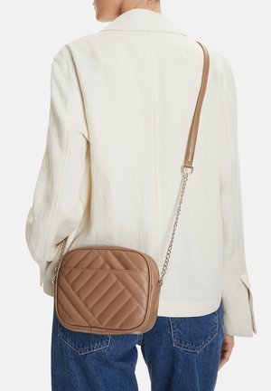 Osoba v krémovém saku a modrých džínách nese malou hnědou prošívanou kabelku přes rameno s řetízkovým popruhem.