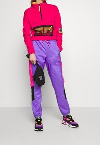 Sudadera de forro polar rosa brillante con un bolsillo de malla, combinada con pantalones de chándal morados y rosas. Incluye una pequeña mochila negra de cintura y zapatillas multicolores.