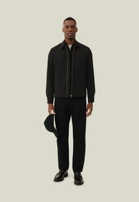 Giacca nera con zip e colletto piatto, indossata sopra una camicia nera e abbinata a pantaloni scuri e scarpe nere. Tiene in mano un cappellino nero.