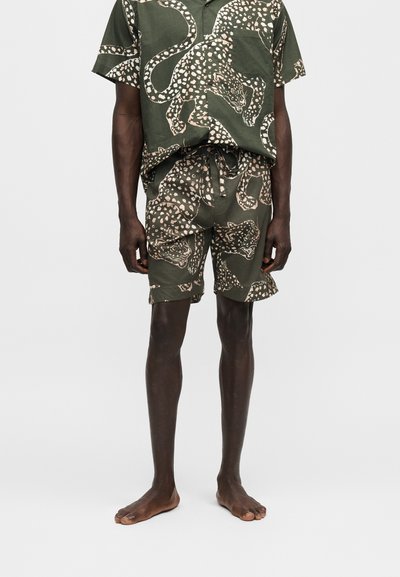 Άνδρας που φοράει ταιριαστό σκούρο πράσινο κοντομάνικο πουκάμισο και σορτς με μεγάλο μπεζ λεοπάρ μοτίβο, όρθιος ξυπόλητος σε απλό φόντο.