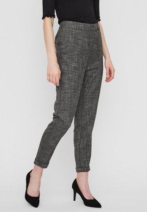 Kobieta ubrana w szare, w kratę, dopasowane spodnie z podwiniętymi nogawkami i czarne buty na wysokim obcasie, stojąca na jednolitym tle.