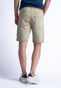 Beige shorts dengan cuff digulung dan saku belakang, dipadukan dengan sepatu sneaker berwarna terang dengan aksen navy. Desain dan tekstur minimalis.