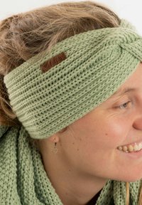 Fascia in maglia verde menta con un design attorcigliato e un piccolo etichetta in pelle, abbinata a una sciarpa in maglia coordinata. Texture morbida e aspetto accogliente.