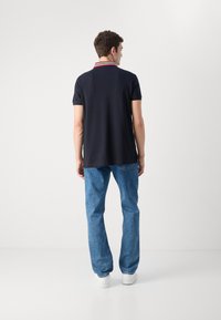 Ναυτικό πόλο μπλουζάκι με κόκκινη και λευκή ριγέ γιακά, συνδυασμένο με ανοιχτό μπλε τζιν παντελόνι. Το μοντέλο απεικονίζεται από το πίσω μέρος, φορώντας λευκά αθλητικά παπούτσια.