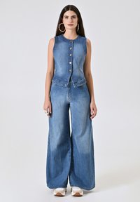 Gilet in denim con design senza maniche e bottoni frontali, abbinato a jeans in denim a gamba larga. Entrambi i capi sono in una tonalità di blu chiaro.