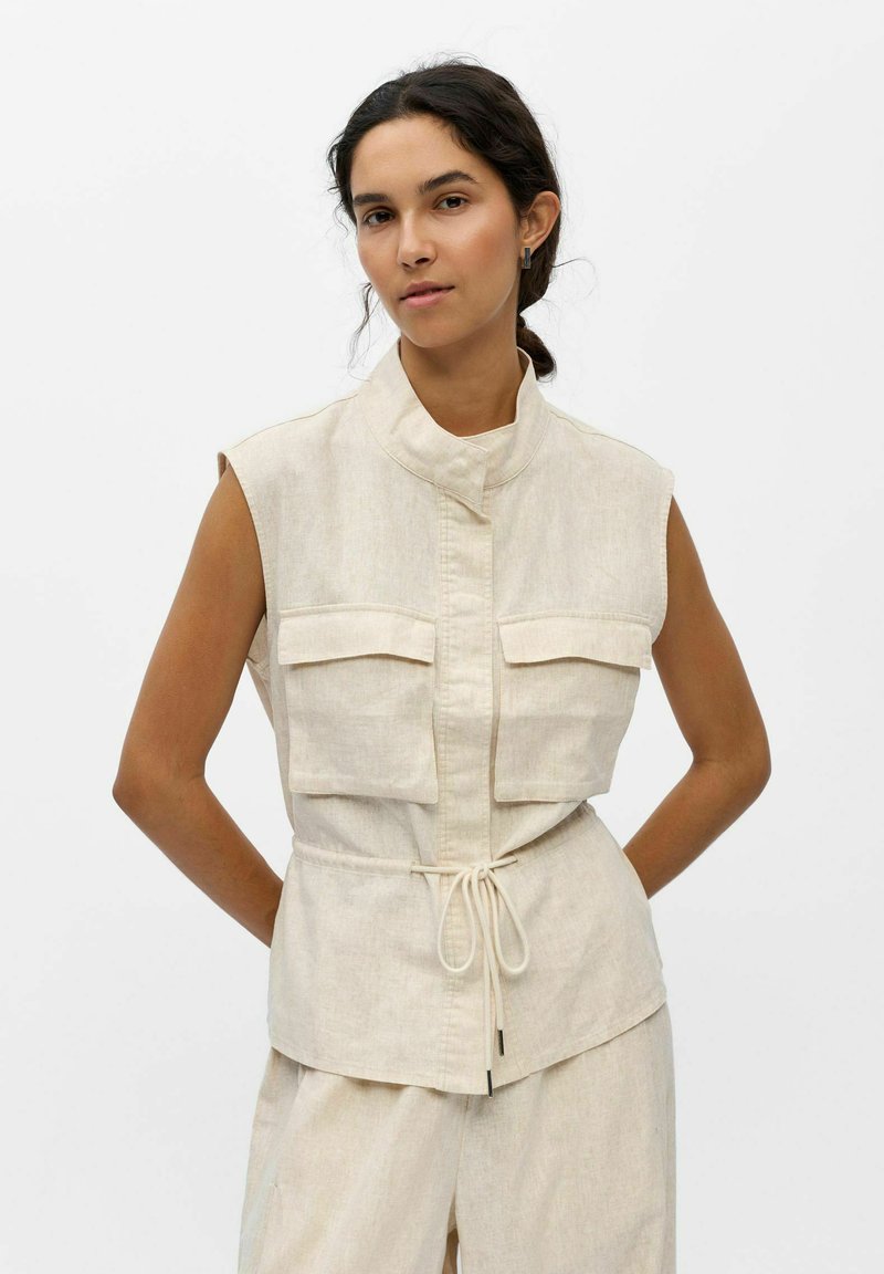 Femme aux cheveux foncés porte un gilet utilitaire beige sans manches avec poches avant et cordon à la taille, sur un pantalon large assorti.