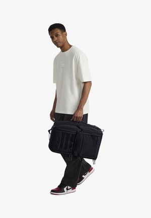 Črna nahrbtnik z več zadrgo predelki, teksturirano blago in tremi navpičnimi trakovi spredaj, ga drži oseba v svetli srajci.