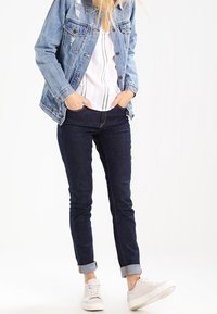 Jeansjacka med ljus tvätt och slitningar, kombinerad med en vit texturerad blus, mörka skinny jeans och vita sneakers.