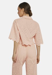 Pink eyelet blouse with short scalloped sleeves, featuring a textured pattern and open holes. The back has a relaxed fit with a collar.