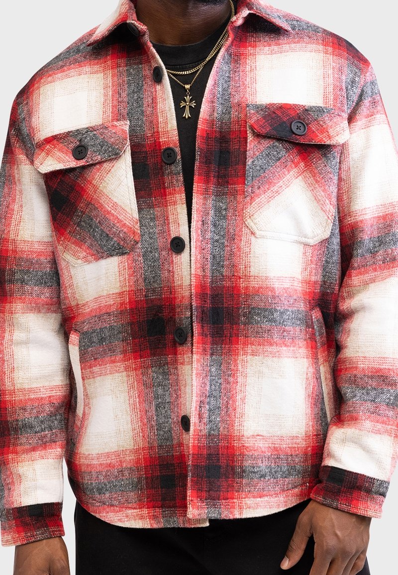 Homme portant une veste à carreaux rouge, blanche et noire boutonnée avec deux poches poitrine et un collier croix en or sur une chemise noire.