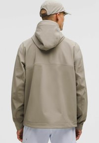 Chaqueta impermeable beige con capucha. Presenta una textura suave, un ajuste ligeramente holgado y una costura horizontal en la parte posterior. Diseño ligero.