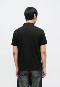 Polo nera in tessuto strutturato, con motivo a rombi e colletto. Maniche corte e orlo dritto, indossata con jeans grigio scuro.