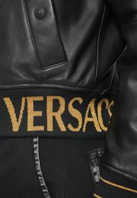 Μαύρο δερμάτινο τζάκετ με έντονη πλεκτή μέση που φέρει χρυσά γράμματα "VERSACE", συμπληρωμένο με αντίθετη ραφή στο ύφασμα.