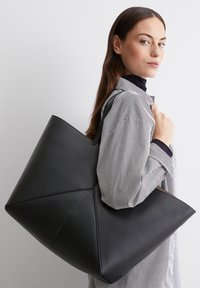 Zwarte leren tote bag met een geometrisch ontwerp, gekenmerkt door scherpe hoeken en een gladde textuur, vastgehouden door een persoon in een gestreepte shirt.