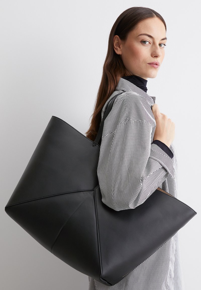 Zwarte leren tote bag met een geometrisch ontwerp, gekenmerkt door scherpe hoeken en een gladde textuur, vastgehouden door een persoon in een gestreepte shirt.