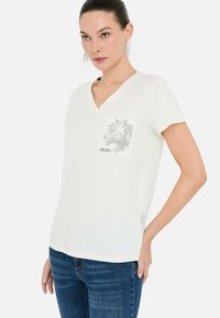 Camiseta blanca de cuello en V hecha de tela suave, con un diseño gráfico de la cabeza de un tigre en el bolsillo izquierdo del pecho, combinada con jeans azules.