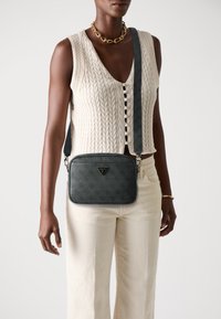 Borsa a spalla nera con texture, logo triangolare, tracolla regolabile e chiusura con zip, abbinata a un top lavorato a maglia crema e pantaloni beige.