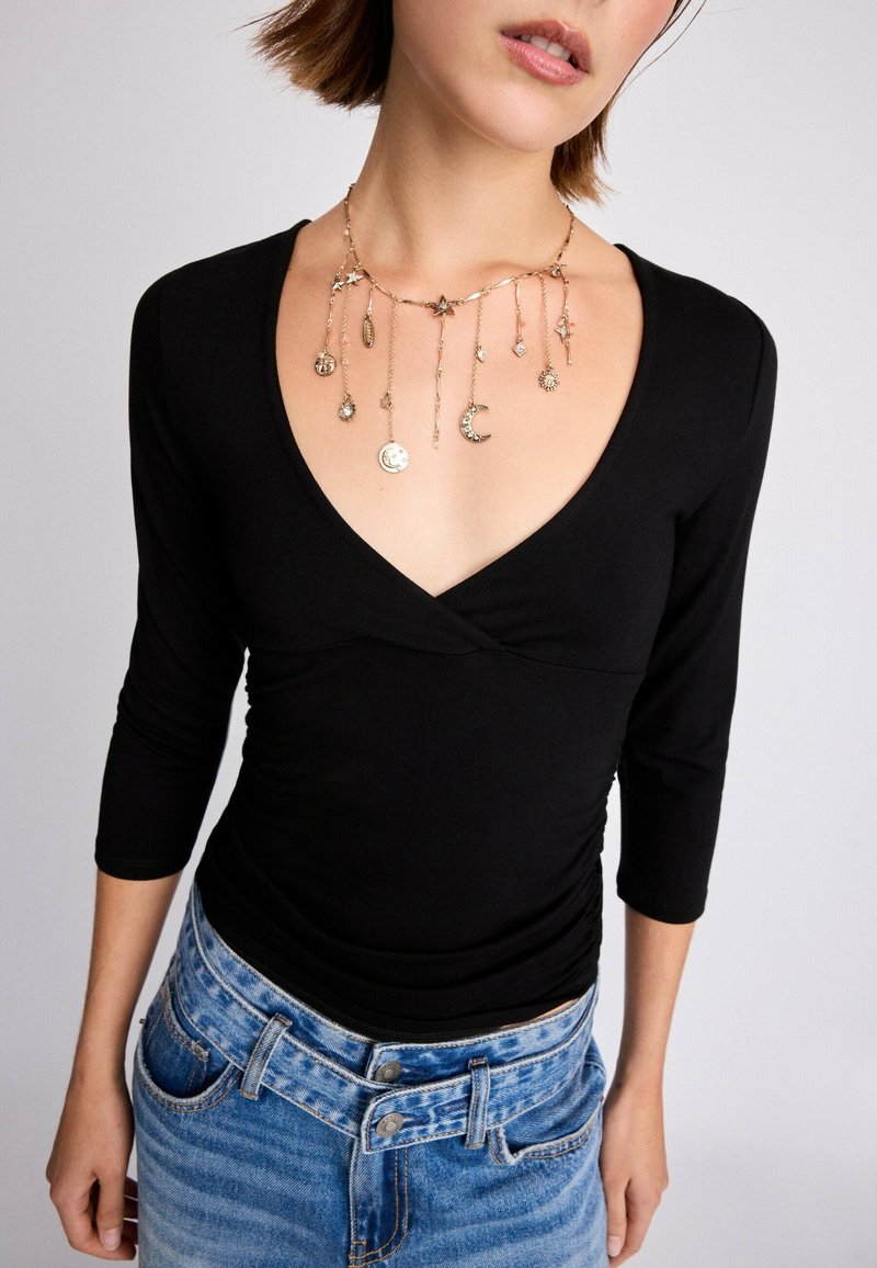 Femme en haut noir col en V et jean bleu porte un collier en or avec des breloques suspendues célestes et étoiles.