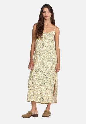 Femme aux longs cheveux bruns portant une robe midi jaune à fleurs sans manches avec une fente latérale et des sabots marron à enfiler.