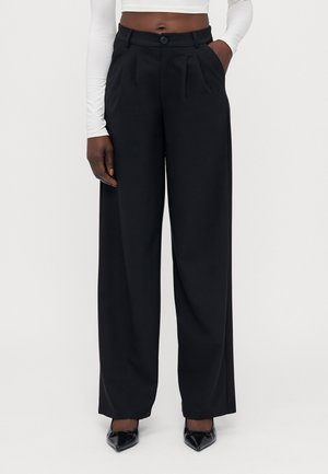 Black wide-leg trousers with front pleats, textured fabric, and a button closure at the waist. Worn with a fitted white top and black heels.