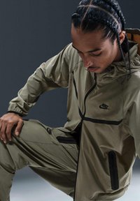 Olivgrön Nike träningsoverall med svarta inslag, blixtlåsfickor och elastiska muddärmar. Lättviktsmaterial, åtsittande design och huva.