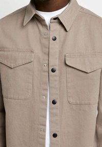 Beżowa koszula dżinsowa z długimi rękawami, czarnymi zatrzaskami, dwoma kieszeniami na piersi oraz fakturowaną powierzchnią tkaniny. Widoczna biała podkoszulka.