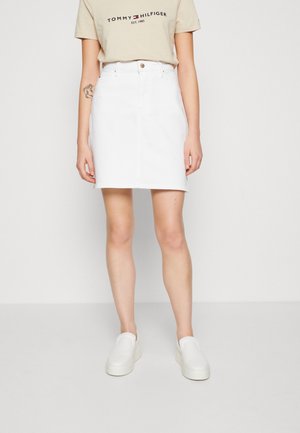 Kobieta ubrana w beżową koszulkę Tommy Hilfiger, białą spódnicę z wysokim stanem, białe buty wsuwane, stojąca na jednolitym białym tle.