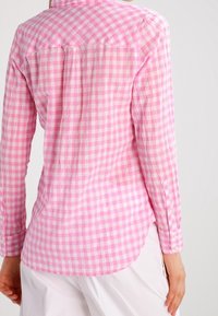 Halenka s dlouhým rukávem v světle růžovém a bílém károvaném vzoru, se stojáčkem, knoflíkovými manžetami a mírně zakřiveným lemem vzadu.