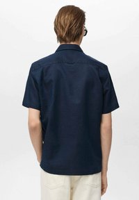 Cămașă navy cu mânecă scurtă, cu guler clasic, design curat pe spate și textură netedă, asociată cu pantaloni de culoare deschisă.