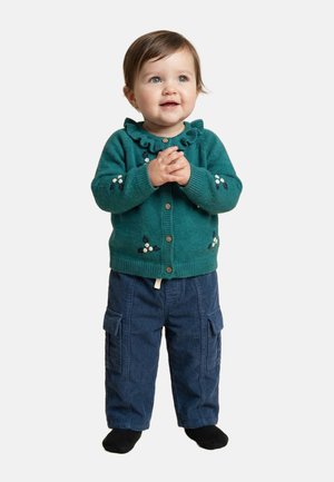 Tout-petit debout, portant un cardigan bleu sarcelle avec broderie florale, un pantalon cargo bleu marine et des chaussettes noires, les mains jointes et regardant vers le haut.